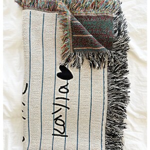 May include: A white woven throw blanket with black stripes and the words "I love you" written in black. The blanket has a fringe border and is folded in half.