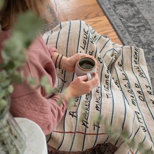 May include: A person sitting on a blanket with blue and white stripes. The blanket has handwritten text on it, including "Love", "Dad", and "More Love". The person is holding a white mug with a green beverage.