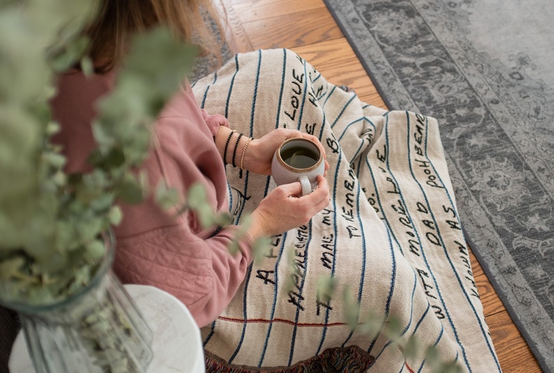 May include: A person sitting on a blanket with blue and white stripes. The blanket has handwritten text on it, including "Love", "Dad", and "More Love". The person is holding a white mug with a green beverage.