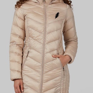 May include: A long beige quilted puffer jacket with a hood and a black lightning bolt logo on the chest. The jacket has a zipper closure and two pockets.