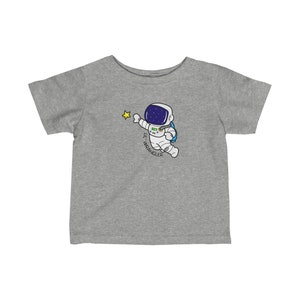 May include: Gray short-sleeved t-shirt with a cartoon astronaut reaching for a yellow star. The astronaut is wearing a white spacesuit with a blue backpack and a helmet with a dark blue visor. The text "Lil Wrangler" is written below the astronaut.