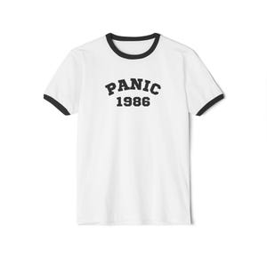 Puede incluir: Camiseta blanca con cuello de canalé negro y "PANIC 1986" impreso en negro en la parte delantera.
