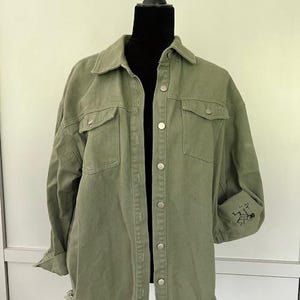May include: A green denim jacket with a button-up front and frayed edges. The jacket has two chest pockets and two front pockets.