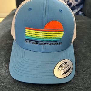 May include: A blue baseball cap with a white mesh back. The front features a colorful sunset design with the text "ONCE MORE I BEAT THE SUNRISE". The cap has a curved brim and a snapback closure.