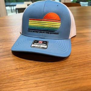 May include: A blue trucker hat with a white mesh back. The front of the hat features an embroidered design of a sunset over colorful stripes, with the text "ONCE MORE I BEAT THE SUNRISE". The hat has a curved brim and an adjustable strap.