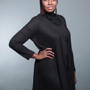 Op de afbeelding: Een vrouw draagt een zwarte hijab en een zwarte gebreide cardigan met een lang, vloeiend ontwerp. Ze staat met haar hand op haar heup en kijkt naar de camera.