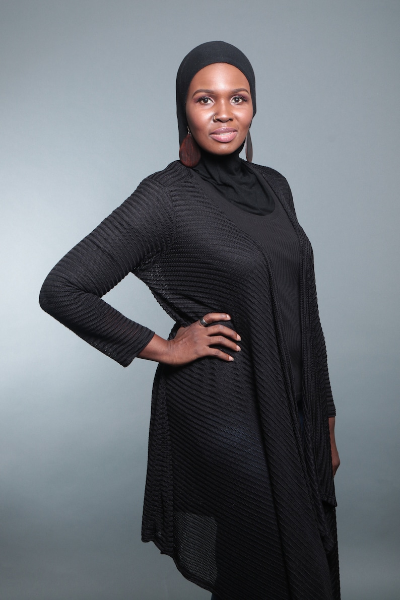 Op de afbeelding: Een vrouw draagt een zwarte hijab en een zwarte gebreide cardigan met een lang, vloeiend ontwerp. Ze staat met haar hand op haar heup en kijkt naar de camera.