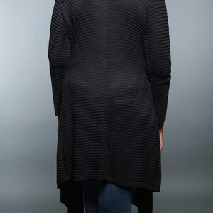 Op de afbeelding: Een zwarte, langarmige cardigan met een geribbelde textuur en een high-low zoom. De cardigan wordt gedragen over een zwart shirt en een blauwe jeans.