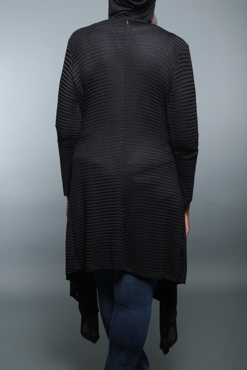 Op de afbeelding: Een zwarte, langarmige cardigan met een geribbelde textuur en een high-low zoom. De cardigan wordt gedragen over een zwart shirt en een blauwe jeans.