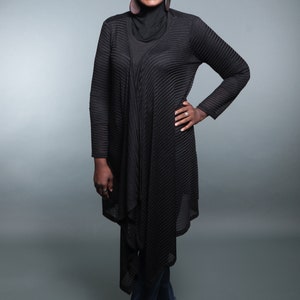 Op de afbeelding: Een vrouw draagt een zwarte hoofddoek en een zwarte, lange, gekreukelde cardigan met een zwarte top eronder. Ze draagt een blauwe jeans en zwarte schoenen.