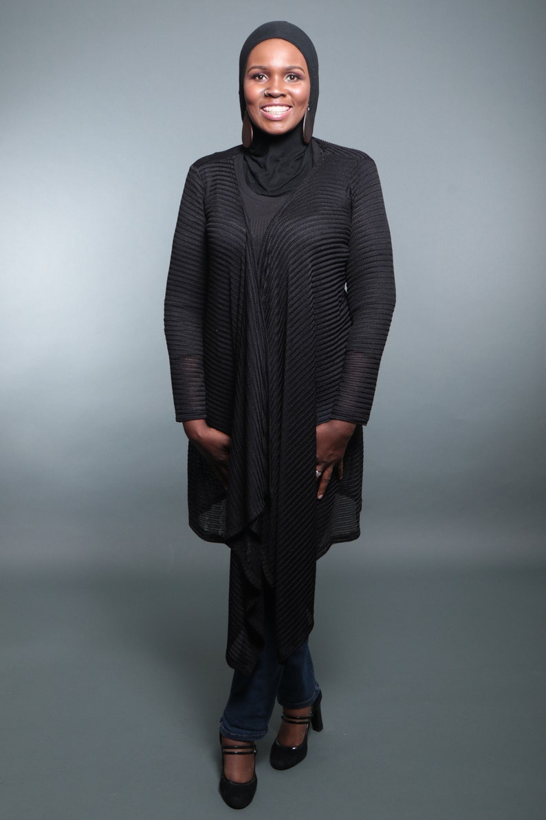 Op de afbeelding: Een vrouw draagt een zwarte hijab, een zwarte gebreide cardigan en een blauwe jeans. Ze glimlacht en kijkt naar de camera.