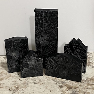 Könnte beinhalten: Fünf schwarze, grabsteinförmige Dekorationen mit weißen Spinnennetzmustern. Die Grabsteine sind in verschiedenen Größen und Formen erhältlich. Ideal für Halloween oder Gothic-Dekorationen.