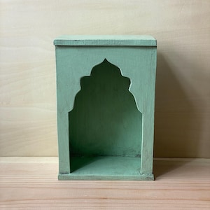 May include: A light green wooden wall shelf featuring a rectangular frame with a decorative arched cutout opening. The structure has a flat top and bottom base, with a matte finish and visible wood grain texture throughout the painted surface.