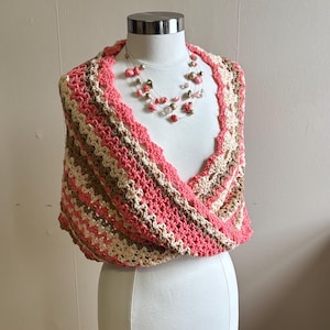 May include: A handmade crochet shawl in shades of pink, beige, and brown, draped over a mannequin. The shawl features a textured stitch pattern and a scalloped edge. A delicate necklace with pink and clear beads is also displayed.