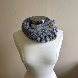 May include: A gray crocheted cowl neck warmer with two wooden buttons. The neck warmer is displayed on a white mannequin. The cowl is made of a thick, textured yarn, perfect for cold weather.