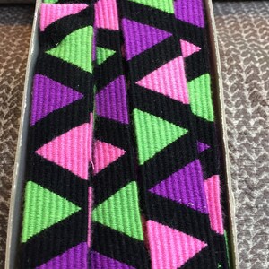 May include: A close-up of a black, pink, green, and purple patterned ribbon. The ribbon is woven with a repeating triangle pattern.