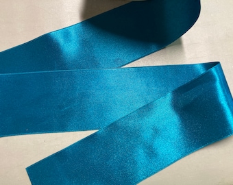 May include: Teal satin ribbon, a smooth and shiny fabric with a subtle sheen. The ribbon is folded in thirds, showcasing its luxurious texture.