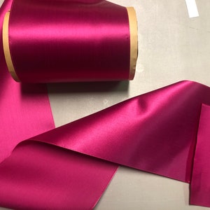 May include: A roll of deep pink satin ribbon with a brown paper core. The ribbon is unfurled and folded on a gray surface.
