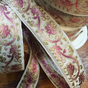 Könnte beinhalten: Nahaufnahme einer Rolle dekorativen Bandes. Das Band hat einen cremefarbenen Hintergrund mit einem sich wiederholenden Muster aus goldenen und rosa floralen Designs. Das Band eignet sich für Bastelarbeiten, Nähen oder Geschenkverpackungen.