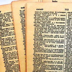 May include: A close-up of several aged dictionary pages, showing definitions. The pages have a yellowed, vintage look. Words such as "alloy", "corsair", and "cosmetic" are visible. The pages are slightly curled, suggesting age.