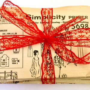 May include: A stack of vintage Simplicity sewing pattern books tied together with a red lace ribbon. The cover of the top book reads "Simplicity Primer Guide for Cutting Sewing - Detail Dressmaking 5698".