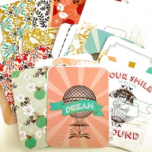 May include: A collection of decorative cards with various patterns and designs. Some cards feature floral and geometric patterns, while others have illustrations of a hot air balloon with the word "DREAM" and a world map with the words "YOUR SMILE makes the World ROUND".