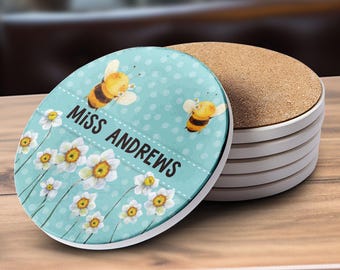 May include: A stack of white ceramic coasters with a light blue top coaster featuring a bee and floral design. The top coaster has the text "MISS ANDREWS" in white. The coasters are round and have a cork backing.