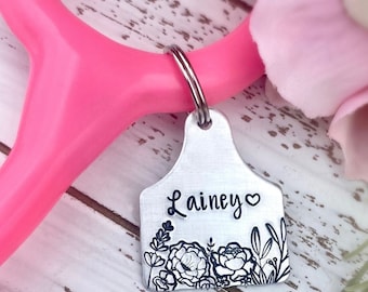 May include: A silver metal tag with the name "Lainey" and a heart symbol, adorned with floral and leaf designs. The tag is attached to a silver keyring and rests against a pink stethoscope. Soft pink and white flowers are in the background.