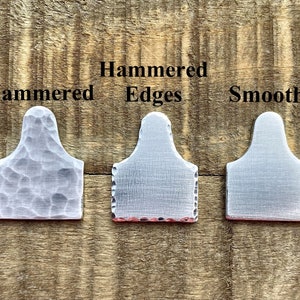 May include: Three metal tags with different surface treatments: hammered, hammered edges, and smooth. The tags are silver-colored and have a rectangular shape with a rounded top. The text "Hammered Edges" and "Smooth" are visible.