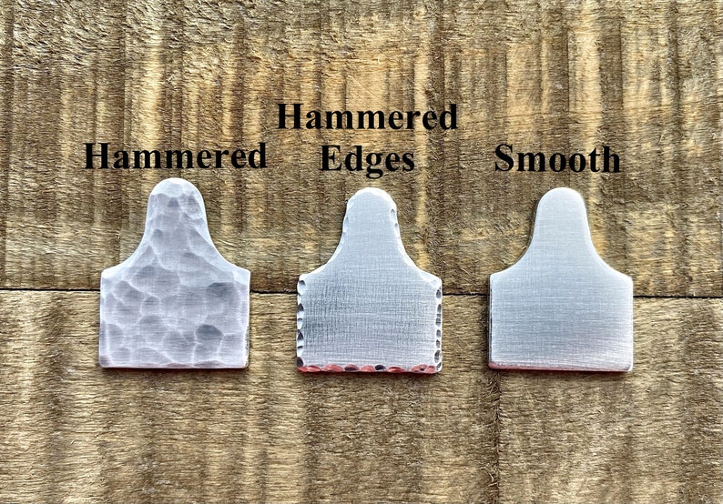 May include: Three metal tags with different surface treatments: hammered, hammered edges, and smooth. The tags are silver-colored and have a rectangular shape with a rounded top. The text "Hammered Edges" and "Smooth" are visible.
