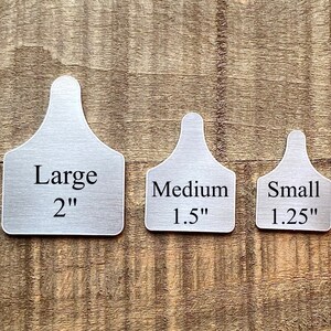 May include: Three metal livestock ear tags in varying sizes: Large (2 inches), Medium (1.5 inches), and Small (1.25 inches). Each tag has a rectangular body with rounded corners and a narrow, curved top for attachment. The tags are displayed on a wooden surface.