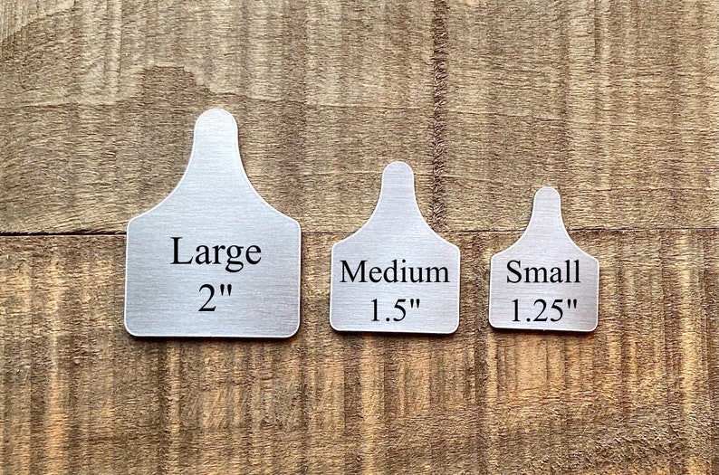 May include: Three metal livestock ear tags in varying sizes: Large (2 inches), Medium (1.5 inches), and Small (1.25 inches). Each tag has a rectangular body with rounded corners and a narrow, curved top for attachment. The tags are displayed on a wooden surface.