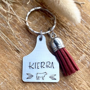 May include: A silver-colored metal keychain with a cow tag design. The tag is stamped with the name "KIERRA", a cow graphic, and arrow symbols. A burgundy tassel hangs from the tag. The keychain is on a silver ring.