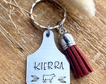 May include: A silver-colored metal keychain with a cow tag design. The tag is stamped with the name "KIERRA", a cow graphic, and arrow symbols. A burgundy tassel hangs from the tag. The keychain is on a silver ring.