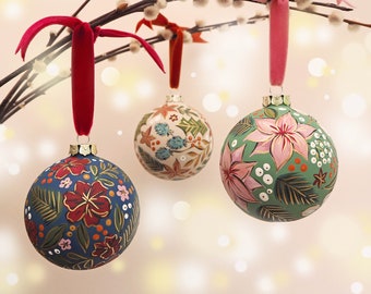 May include: Three hand-painted Christmas ornaments with floral designs. The ornaments are green, blue, and white with red and pink flowers. The ornaments are hanging from a branch with white fuzzy buds.