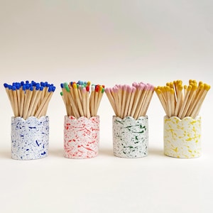 May include: Four decorative match holders with scalloped edges, each filled with matches. The holders are white with colourful paint splatters in blue, red, green, and yellow. The matches have coloured tips.