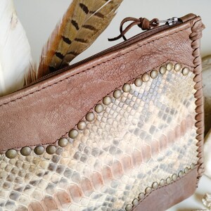 May include: A brown leather zippered pouch with a snakeskin pattern and metal studs. The pouch is decorated with a brown feather.