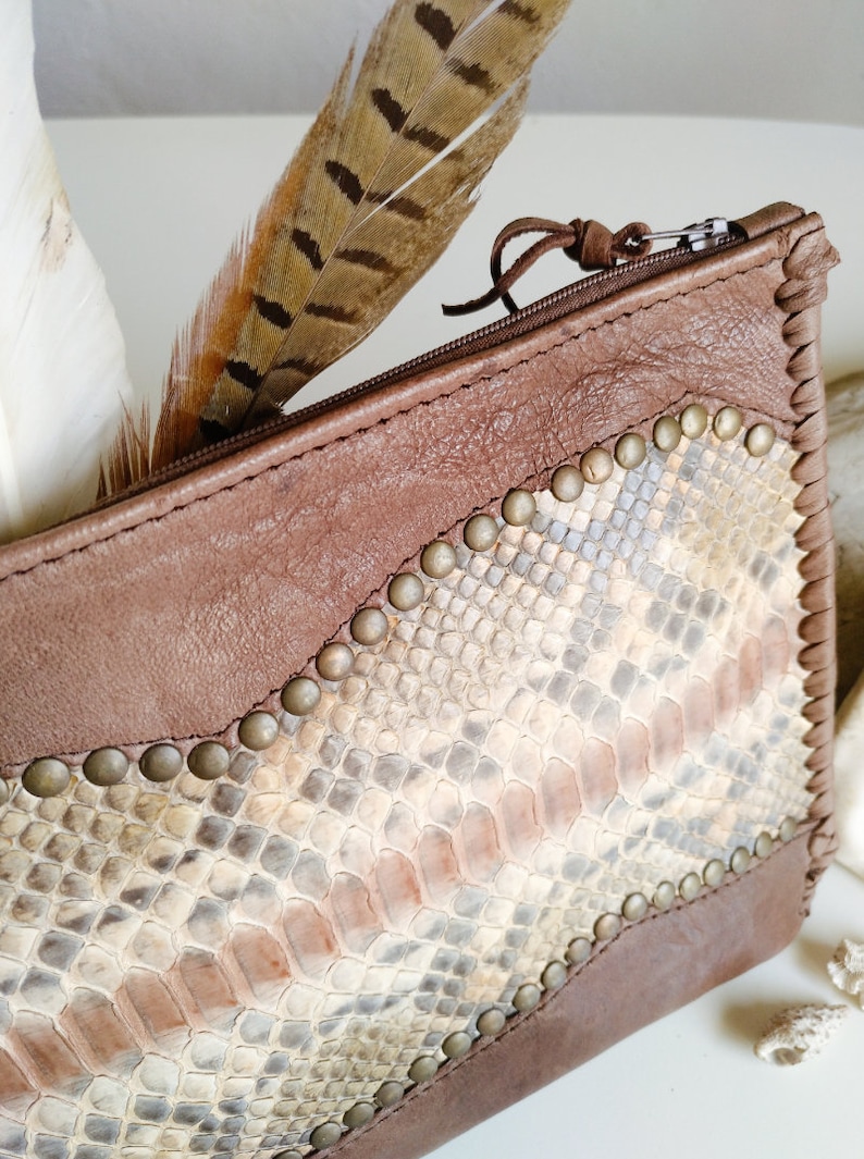 May include: A brown leather zippered pouch with a snakeskin pattern and metal studs. The pouch is decorated with a brown feather.