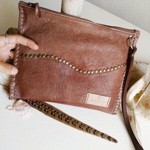 May include: A brown leather clutch with a zipper closure and a long strap. The clutch has a decorative row of metal studs along the top edge and a small leather label with the text "Handmade" on the front.