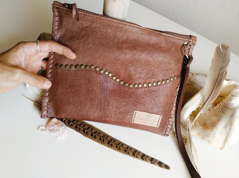 May include: A brown leather clutch with a zipper closure and a long strap. The clutch has a decorative row of metal studs along the top edge and a small leather label with the text "Handmade" on the front.