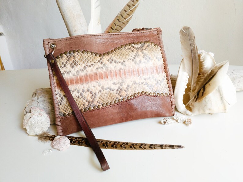 May include: A brown leather clutch with a snakeskin pattern and a long strap. The clutch has a flap closure and is decorated with metal studs.
