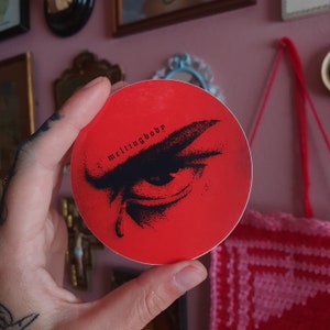 Puede incluir: Una pegatina redonda roja con una ilustración en blanco y negro de un ojo con el ceño fruncido. El texto "meltingbody" está impreso en blanco en la pegatina.