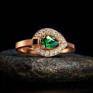 Puede incluir: Un anillo de oro rosa con una gema verde esmeralda en forma de pera, rodeada por un halo de peque&ntilde;as gemas claras. El anillo est&aacute; colocado sobre una piedra oscura y texturizada sobre un fondo negro.