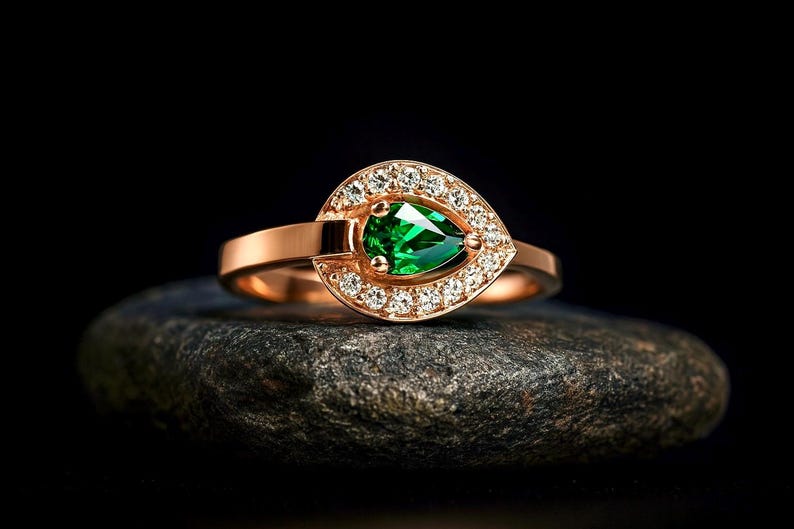 Puede incluir: Un anillo de oro rosa con una gema verde esmeralda en forma de pera, rodeada por un halo de peque&ntilde;as gemas claras. El anillo est&aacute; colocado sobre una piedra oscura y texturizada sobre un fondo negro.