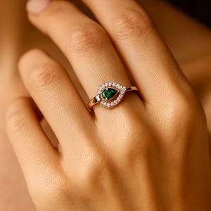 Puede incluir: Anillo de oro rosa con una piedra preciosa verde esmeralda en forma de pera, rodeada de peque&ntilde;as piedras preciosas claras. El anillo, engarzado en una banda delicada, presenta un dise&ntilde;o cl&aacute;sico y elegante. El anillo se muestra en una mano.
