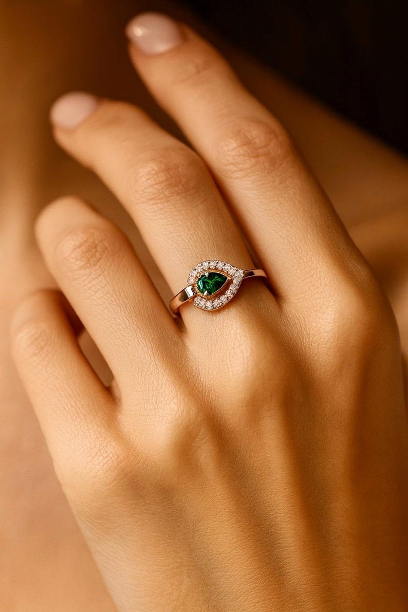 Puede incluir: Anillo de oro rosa con una piedra preciosa verde esmeralda en forma de pera, rodeada de peque&ntilde;as piedras preciosas claras. El anillo, engarzado en una banda delicada, presenta un dise&ntilde;o cl&aacute;sico y elegante. El anillo se muestra en una mano.