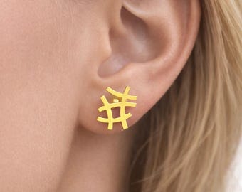 May include: A close-up of a gold-colored stud earring in the shape of an abstract design. The earring is worn on an ear, and the design appears to be a series of intersecting lines. The earring is a bright, shiny gold color.