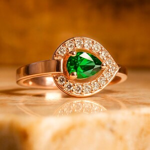 Puede incluir: Un anillo de oro rosa con una gema verde esmeralda en forma de pera. La gema est&aacute; rodeada por un halo de peque&ntilde;os diamantes brillantes. El anillo est&aacute; sobre una superficie clara.