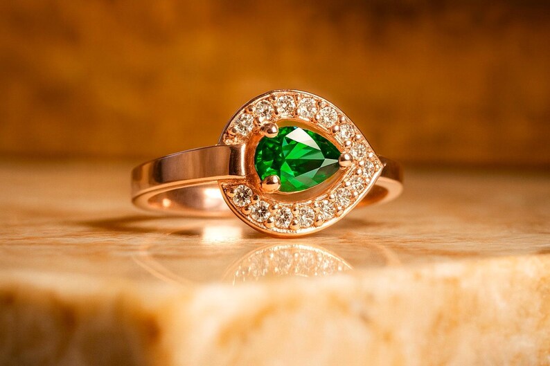 Puede incluir: Un anillo de oro rosa con una gema verde esmeralda en forma de pera. La gema est&aacute; rodeada por un halo de peque&ntilde;os diamantes brillantes. El anillo est&aacute; sobre una superficie clara.