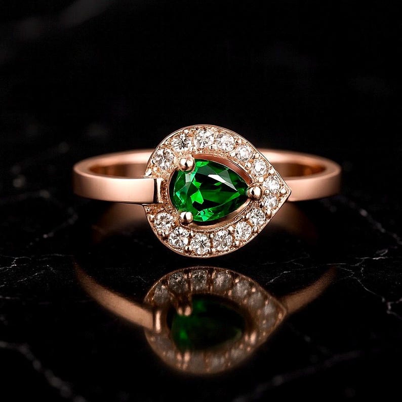 Puede incluir: Anillo de oro rosa con una gema verde vibrante en forma de pera, rodeada de peque&ntilde;os diamantes blancos brillantes. La banda del anillo es de metal pulido, sobre un fondo oscuro. Joya elegante.